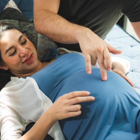 Pregnant Asia woman and husband holding headphones on tummy and playing music guitar to her baby together